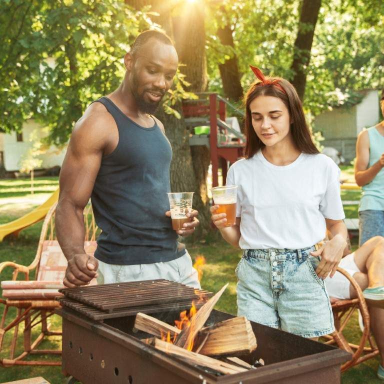 Group of happy friends having beer and barbecue party at sunny day. Resting together outdoor in a forest glade or backyard. Celebrating and relaxing, laughting. Summer lifestyle, friendship concept.